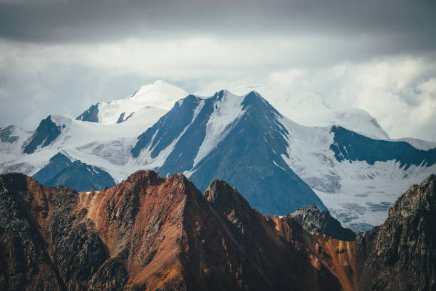 Atmospheric mountains landscape with great snowy top behind colorful brown red orange rocky wall under cloudy sky. Dramatic highland scenery with giant glacier and big vivid brown red orange mountain. paisaje atmosférico de montañas con gran cima nevada detrás de una colorida pared rocosa marrón rojo naranja bajo un cielo nublado. espectacular paisaje de tierras altas con glaciar gigante y gran montaña de color marrón rojo anaranjado vivo.