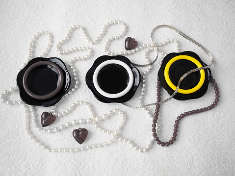 Flat lay. A beautiful composition of glass black dishes, plastic jewelry, bracelets, beads, hearts on a white background. Women's baubles for February 14, Valentine's Day. Still life.