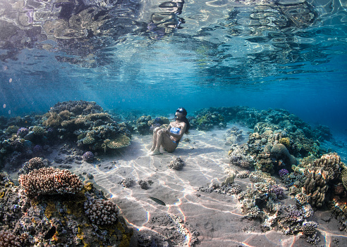 Japanese Underwater model sitting in the sand between coral reef formations in the Red Sea of Egypt while holding her breath