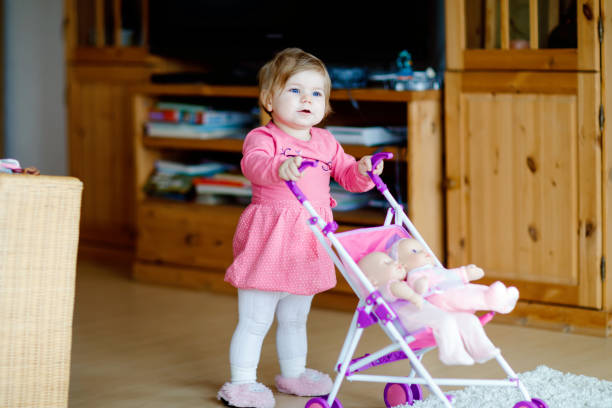 かわいい愛らしい女の赤ちゃんは、最初のステップを作り、人形のキャリッジでプッシュします。自宅でおもちゃでベビーカーを押す美しい幼児の子供。2つの人形を持つ幸せな子供 - baby doll dress ストックフォトと画像