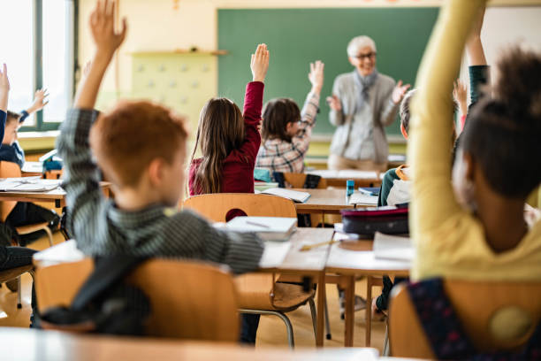 istock Back view of students raising their hands to answer the teacher's question on a class. 1347832895