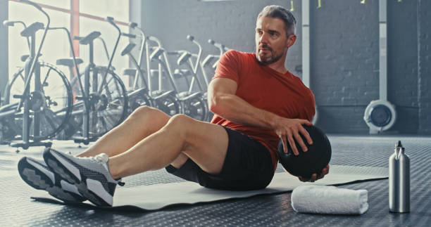 photo d’un bel homme mûr utilisant un ballon de médecine pendant son entraînement dans le gymnase - action male only men mature men photos et images de collection