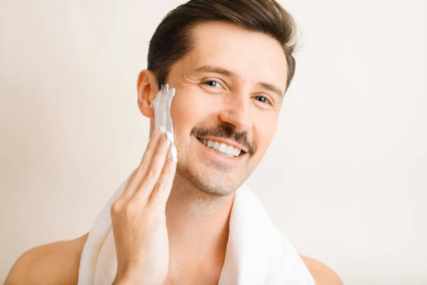 istock Handsome smiling unshaven young man applying moisturizing shaving foam on face and looking at camera. Portrait of happy man with towel on shoulders performing daily self-care, beauty routine indoors 1347837061