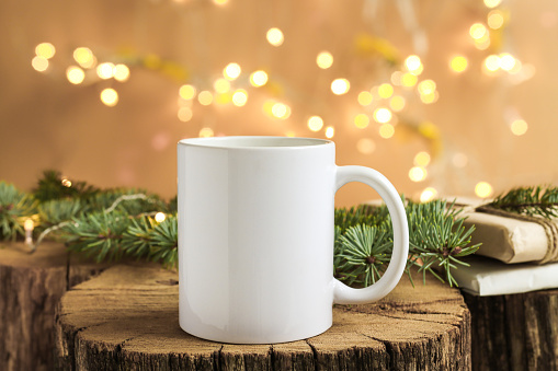 White mug on a wooden background with bright lights in defocus and gifts in the background. Close-up of a ceramic cup for advertising and design for New Year and Christmas