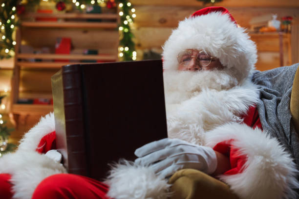 Portrait of Santa reading christmas stories while sitting in a chair Portrait of Santa Claus sitting relaxed in an armchair and reading a fascinating book in his cozy log house animator photos stock pictures, royalty-free photos & images