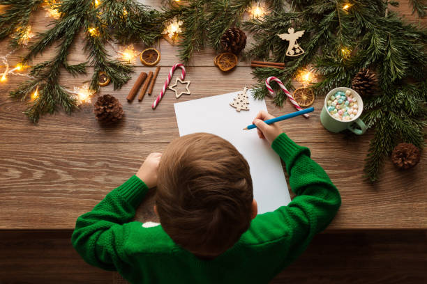 dziecko piszące świąteczny list do świętego mikołaja. widok z góry dziecka siedzącego przy drewnianym stole z lampkami do dekoracji drzew bożoświetnych i pustą białą listą życzeń - christmas christmas ornament human hand decoration zdjęcia i obrazy z banku zdjęć