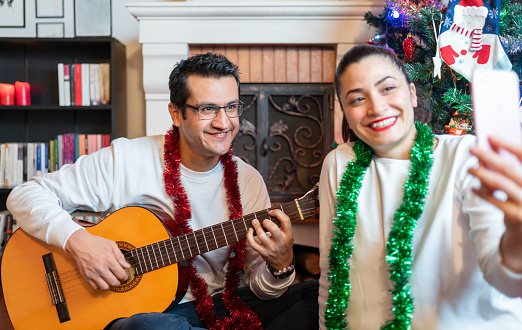 Couple celebrating Christmas at home, playing guitar in front of fireplace and having video call with friends Stock Picture