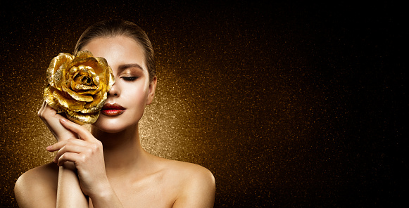 Woman Beauty Perfect glowing Skin Makeup. Fashion Model holding Golden Rose Flower over Face and covering Closed Eye. Artistic glittering Dark Background with Copy Space