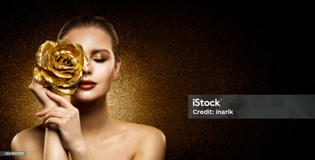 Woman Beauty Perfect glowing Skin Makeup. Fashion Model holding Golden Rose Flower over Face and covering Closed Eye. Artistic glittering Dark Background with Copy Space Woman Beauty Perfect glowing Skin Makeup. Fashion Model holding Golden Rose Flower over Face and covering Closed Eye. Artistic glittering Dark Studio Background with Copy Space Women Stock Photo