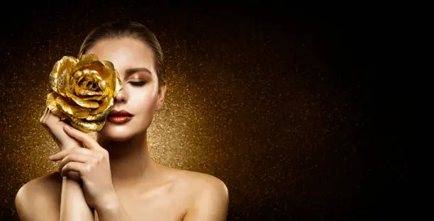 Woman Beauty Perfect glowing Skin Makeup. Fashion Model holding Golden Rose Flower over Face and covering Closed Eye. Artistic glittering Dark Studio Background with Copy Space