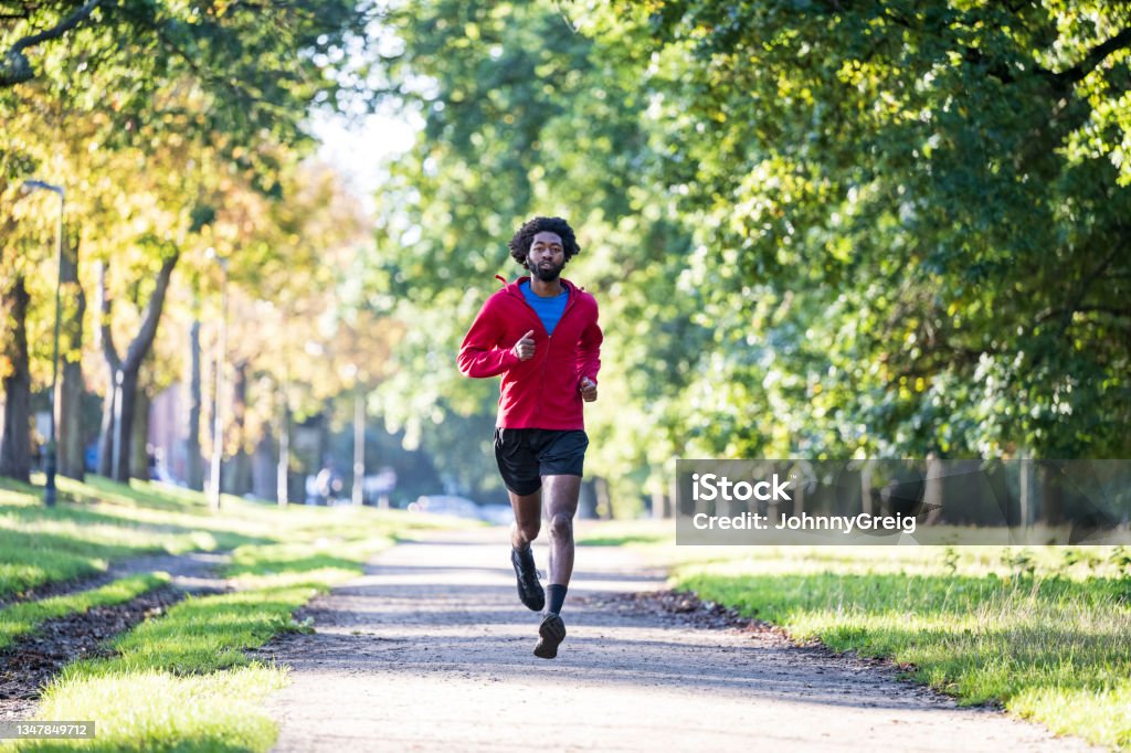 Fit young man enjoying daily run through urban tree area Full length front view of Black athlete in workout attire approaching camera on footpath in early autumn. Running Stock Photo