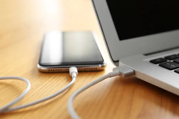 Photo of Modern smartphone charging from laptop on wooden table, closeup