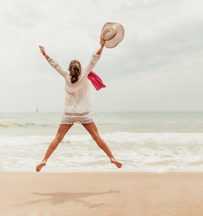 woman jumping on the shore of the beach with hat in hand. she stays slim and jumps with ease. she is happy celebrating something. post pandemic vacation. vaccine.