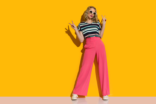Cool young woman in loose pants and sunglasses is standing on front of yellow wall and showing peace hand sign. Full length shot.