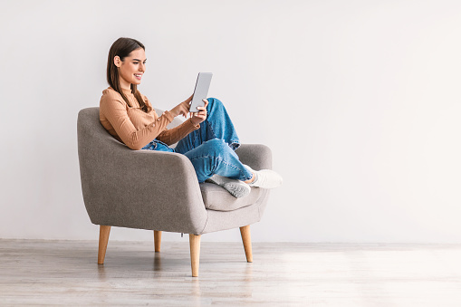 Happy millennial lady using tablet pc, studying or working remotely, having online conference, sitting in armchair against white studio wall, copy space. Young woman with touch pad checking new app