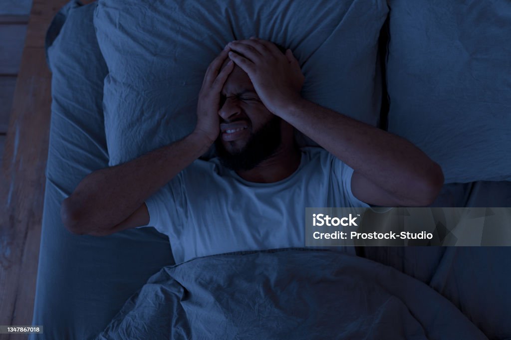 Black man suffering from headache or migraine at night Sleeping Problem. Overhead above top view of stressed African American guy lying alone in bed touching grabbing head feeling depressed, suffering from insomnia or mental problems after breakup Insomnia Stock Photo
