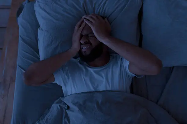 Sleeping Problem. Overhead above top view of stressed African American guy lying alone in bed touching grabbing head feeling depressed, suffering from insomnia or mental problems after breakup