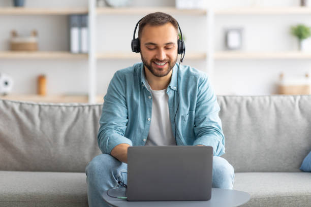 istock Smiling young Caucasian guy in headset making video call on laptop at home, talking to webcam 1347867039