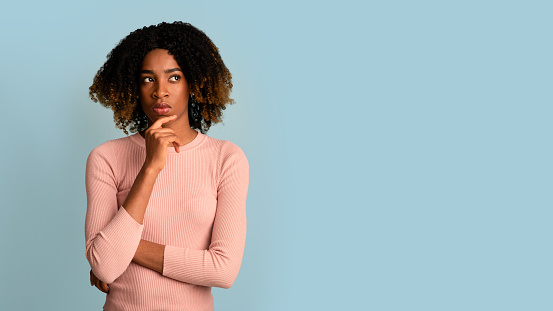 Portrait Of Pensive Black Woman Touching Chin And Looking Aside At Copy Space, Thoughtful African Female Thinking About Offer, Considering Options While Standing Over Blue Background, Panorama