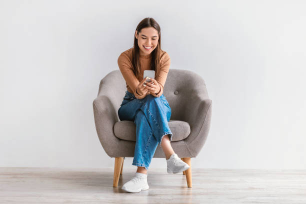 Happy Caucasian woman using cellphone, chatting on internet, working or studying online, sitting in armchair Happy Caucasian woman using cellphone, chatting on internet, working or studying online, sitting in armchair against white studio wall, full length. Cheery young lady watching video on mobile phone one woman only stock pictures, royalty-free photos & images