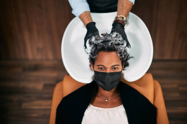 Above view of black woman wearing face mask while getting her hair washed at hairdresser's. High angle view of African American woman washing hair at hair salon and wearing face mask due to COVID-19 pandemic. black woman washing hair stock pictures, royalty-free photos & images