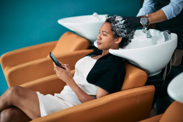 Black woman texting on mobile phone while washing hair at hairdresser's. African American woman using smart phone while getting her hair washed at hair salon. washing hair stock pictures, royalty-free photos & images