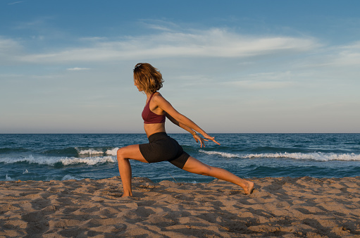 Side view of slim female in activewear doing Ashta Chandrasana pose on sandy beach near waving sea while practicing yoga at sunrise