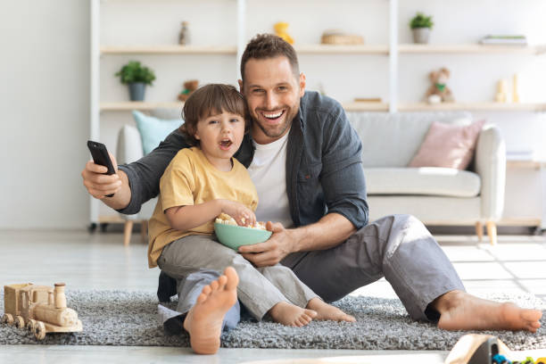 familienzeitvertreib. emotionaler junger vater, der mit seinem kleinen sohn filme im fernsehen sieht und mit popcorn auf dem boden sitzt - soccer child indoors little boys stock-fotos und bilder