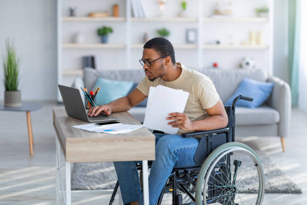 istock Paraplegic black guy in wheelchair with documents using laptop to work online from home 1347877866