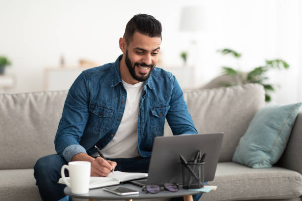 istock Happy young Arab guy working or studying on laptop computer, taking notes, having business meeting indoors 1347878576