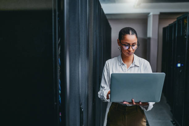 scatto di una giovane donna che usa un laptop mentre lavora in una sala server - it professional foto e immagini stock