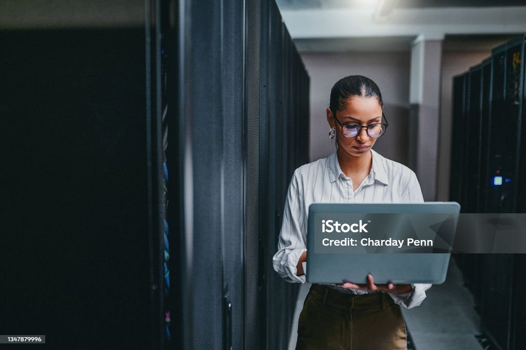 Shot of a young woman using a laptop while working in a server room It takes a technical mind to figure it out Technology Stock Photo