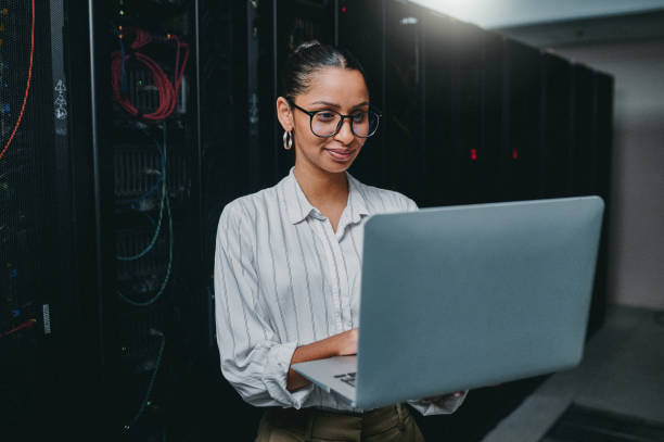 istock Shot of a young woman using a laptop while working in a server room 1347880003