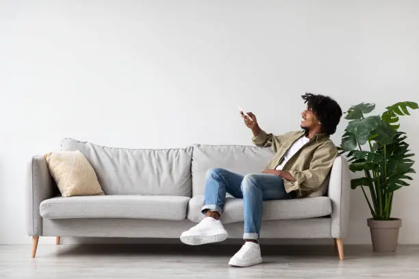 Photo of Cheerful Black Guy Holding Remote Controller, Opening Air Conditioner At Home