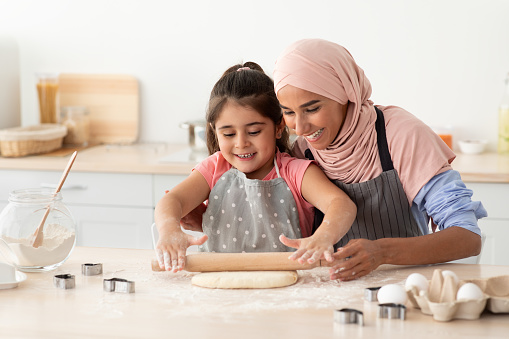 Muslim Mother And Little Daughter Rolling Up Dough On Table While Baking In Kitchen Together, Cute Small Girl And Islamic Mom In Hijab Having Fun At Home, Enjoying Preparing Homemade Pastry