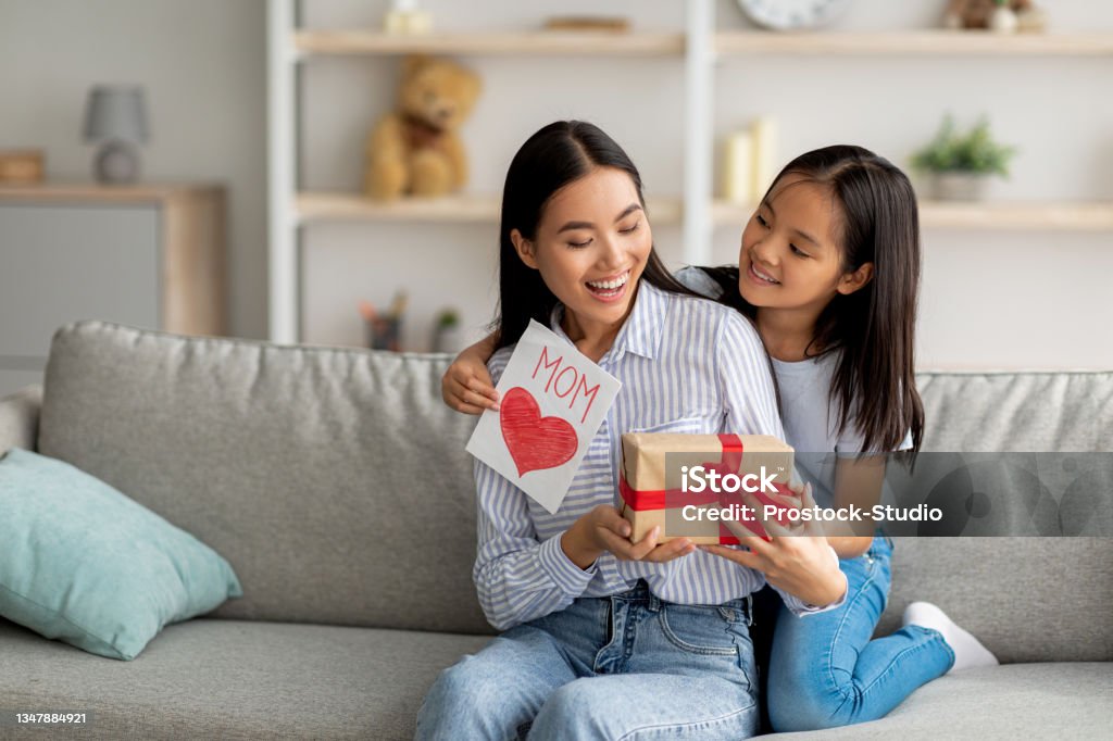 Congratulations. Loving asian daughter greeting surprised mom with Mother's Day, giving her handmade card and gift box Congratulations. Loving asian daughter greeting surprised mom with Mother's Day, giving her handmade card and gift box, sitting on sofa at home interior, free space Mother's Day Stock Photo