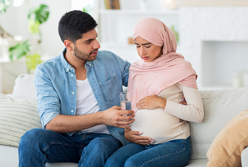 Care and support during pregnancy. Muslim expecting woman feeling sick, man giving her glass of water, siting on sofa at home interior, copy space. Arab husband supporting his woman during pregnancy