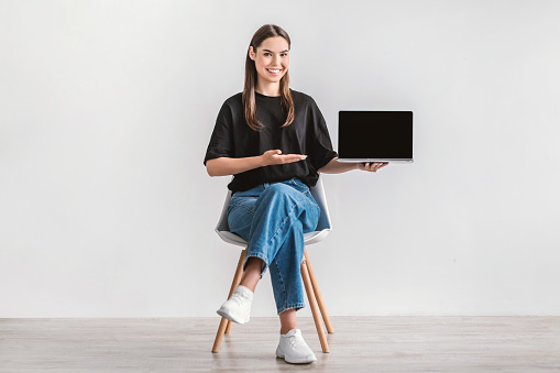 Positive young woman showing laptop pc with empty screen, sitting on chair against white studio wall, mockup for website. Millennial lady presenting portable computer with ad template