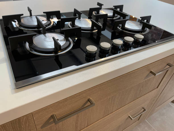 istock Close-up image of modern kitchen 5 burner gas hob stove, five ring kitchen island with under counter storage draws, central wok ring, white stone Corian countertop and black glass surface, row of dials and knobs, elevated view, focus on foreground 1347888409