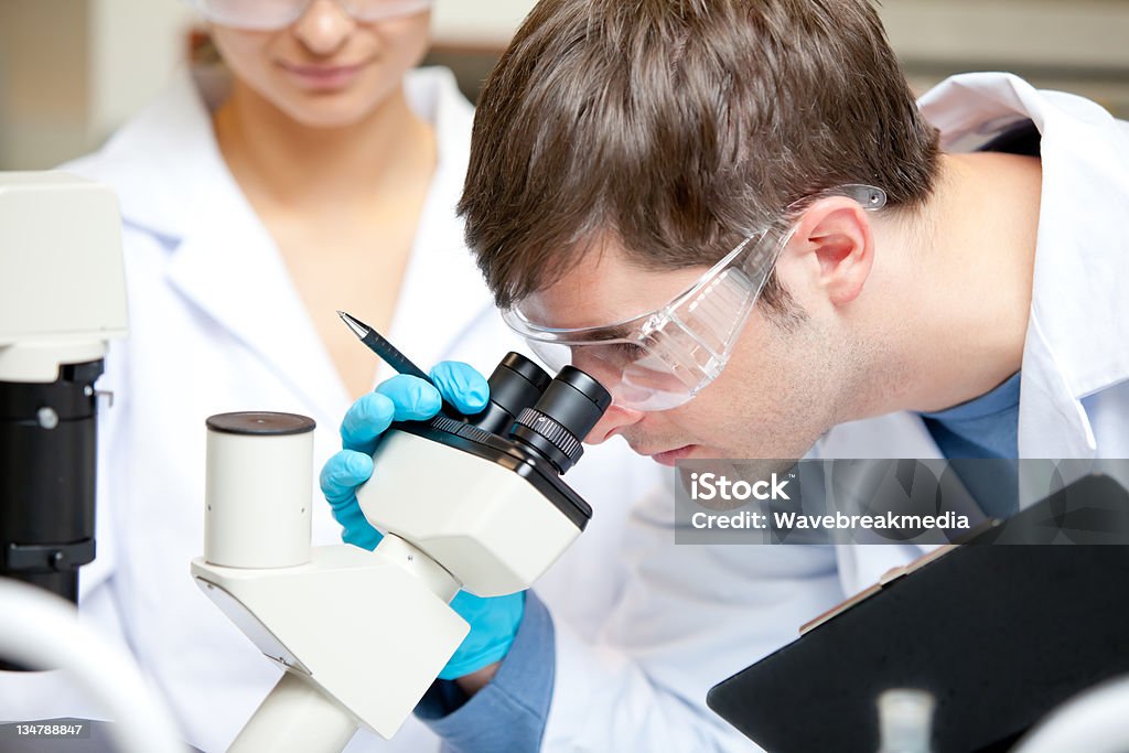 Caucasian male scientist holding pen and clipboard looking throu Caucasian male scientist holding pen and clipboard looking through a microscope in his laboratory Adult Stock Photo