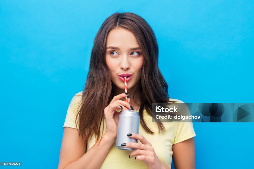 young woman or teenage girl drinking soda from can people concept - smiling young woman or teenage girl in yellow t-shirt drinking soda from can through paper straw over bright blue background Drinking Stock Photo