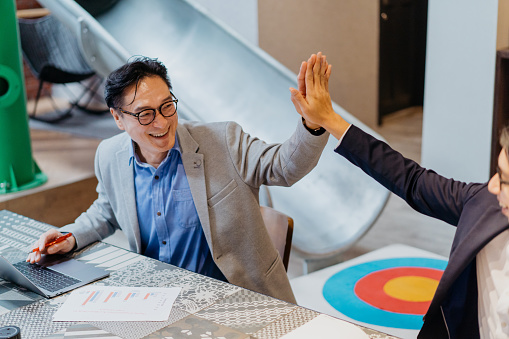 Image of two excited asian businessmen celebrating success and giving each others a high five in coworking office