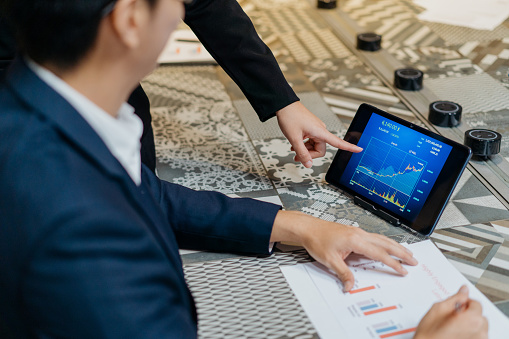 Image of two Asian business people checking stock market chart on digital tablet. Fund manager looking at stock market chart on digital tablet.