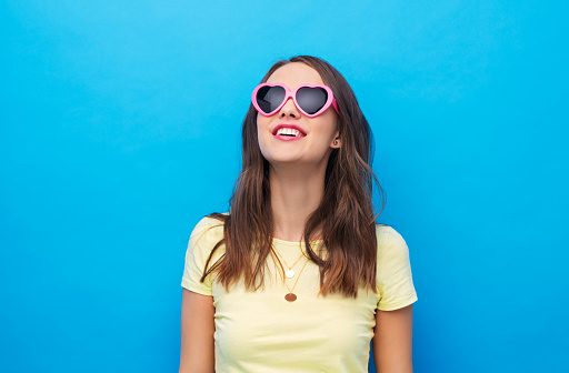 summer, valentine's day and people concept - smiling young woman or teenage girl in yellow t-shirt and heart-shaped sunglasses over bright blue background