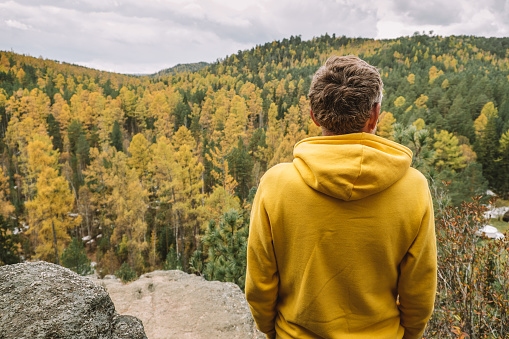 A man in a yellow hoodie stands on top of a mountain, admiring the magnificent landscape of the autumn forest from above. Selective focus. Sustainable environment. People from behind. Close-up.