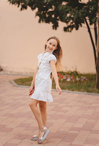 A cute smiling little girl in a summer white dress and gray lace-up shoes posing outdoors. A child on a walk in the park on a warm summer day. Portrait of a girl. Selective focus, defocus. Toning.
