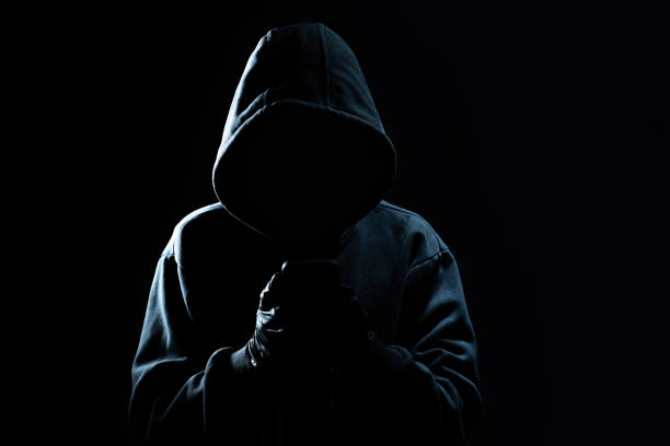 istock Hacker Using Smartphone. Men in black clothes with hidden face looks at smartphone screen on black background 1347894709