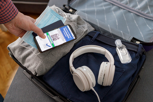 Male hands placing protective mask, passport and mobile phone with International Certificate of Vaccination next to a headphones and hand sanitizer in a suitcase