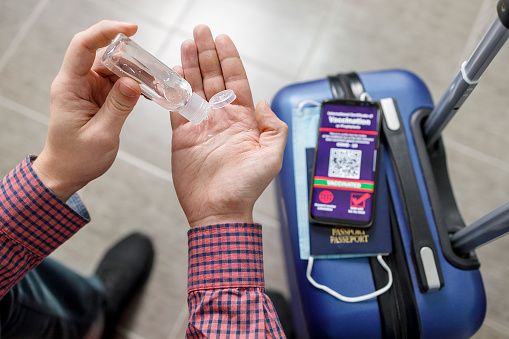Male hands holding a bottle of sanitizer when sitting by a suitcase with mask, passport and vaccination certificate on it
