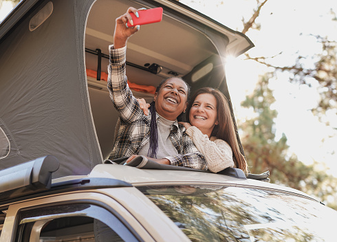 Multiracial senior friends taking a selfie on mini van camper roof using mobile phone